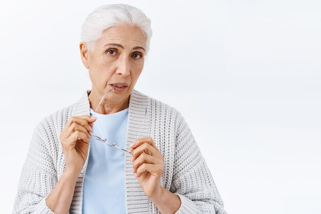 Curious, thoughtful cute senior woman with grey combed neat haircut, take-off glasses, biting rim of eyewear and gaze camera indecisive, making choice