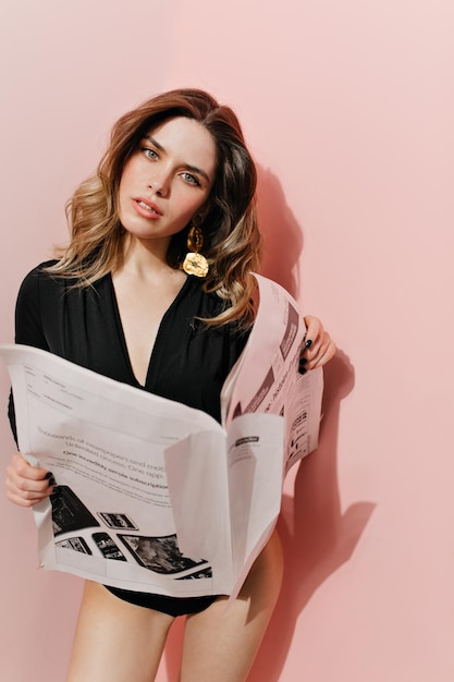 Curious woman holding newspaper Studio shot of caucasian girl in black bodysuit isolated on pink background