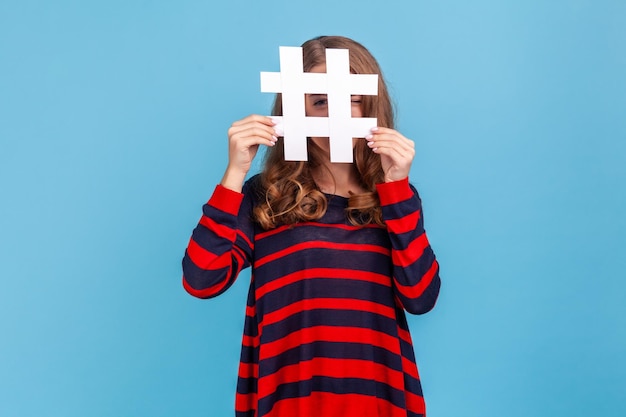 Curious woman wearing striped casual style sweater looking at camera through large white hashtag symbol with prying eye interesting web content Indoor studio shot isolated on blue background