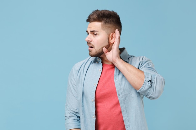 Curious young bearded guy 20s in casual shirt posing isolated on pastel blue wall background studio portrait. People emotions lifestyle concept. Mock up copy space. Try to hear you with hand near ear.