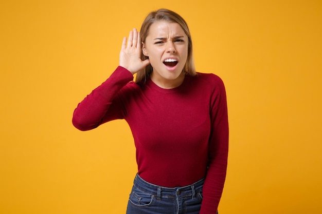 Curious young blonde woman girl in casual clothes posing isolated on yellow orange background studio portrait. People emotions lifestyle concept. Mock up copy space Try to hear you with hand near ear.