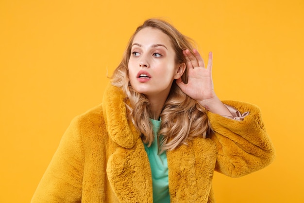 Curious young blonde woman girl in yellow fur coat posing isolated on orange wall background studio portrait. People emotions lifestyle concept. Mock up copy space. Try to hear you with hand near ear.