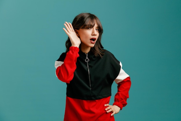 Curious young caucasian woman keeping hand on waist and another hand near ear looking at camera listening isolated on blue background