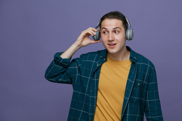 Curious young male student wearing headphones opening one ear looking at side trying to hear through loud sound of music isolated on purple background