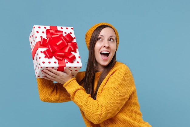 Curious young woman girl in sweater hat posing isolated on blue background. Valentine's Day Women's Day, birthday, holiday concept. Mock up copy space. Hold white red present box with gift ribbon bow.