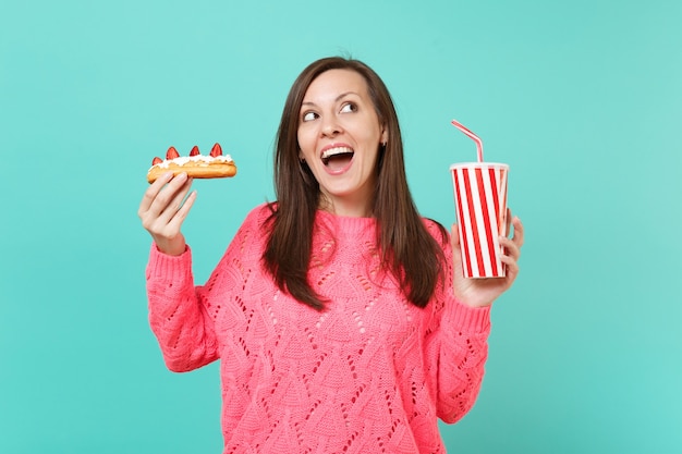 Curious young woman in knitted pink sweater looking up hold in hands eclair cake plastic cup of cola or soda isolated on blue background, studio portrait. People lifestyle concept. Mock up copy space.