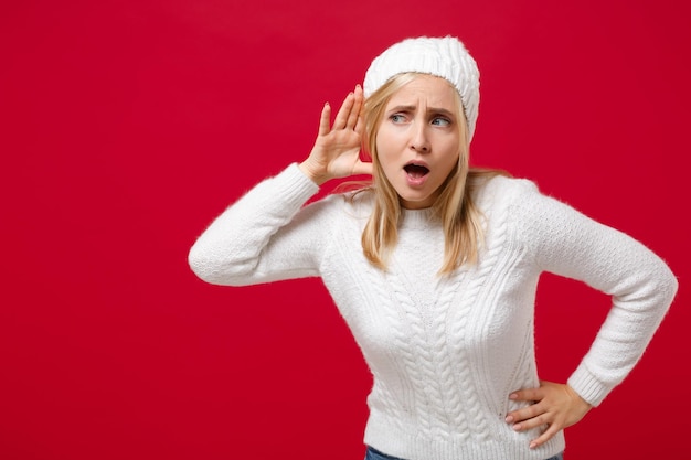 Foto giovane donna curiosa in maglione bianco, cappello isolato su sfondo rosso in studio. stile di vita sano, emozioni della gente, concetto di stagione fredda. simula lo spazio della copia. cerca di sentirti con la mano vicino all'orecchio.