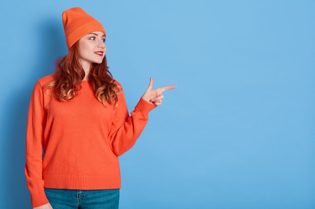 Curious young woman with curly red hair, points aside, dressed in casual sweater, hat and jeans