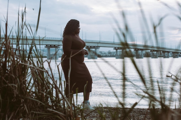 Curly African American plump woman walking near sea coast bridge Plants foreground Dusk sky horizon in summer weekend