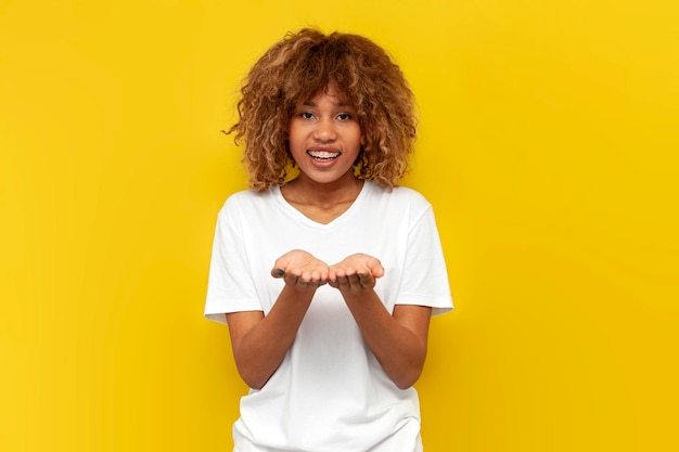 curly american girl with braces holds empty palms in front of her on yellow isolated background