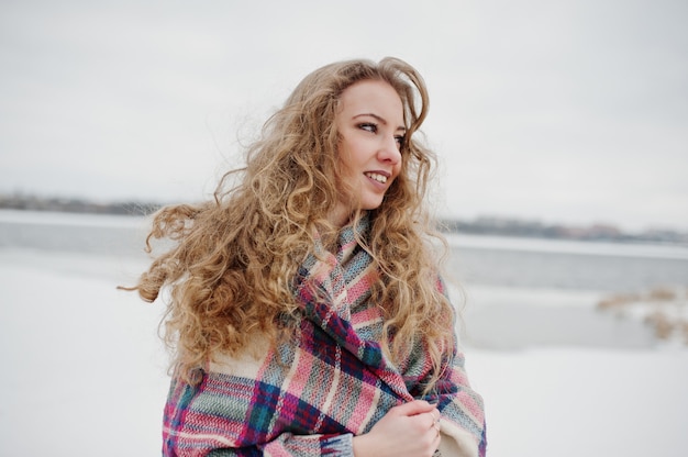 Curly blonde girl in checkered plaid against frozen lake at winter day.