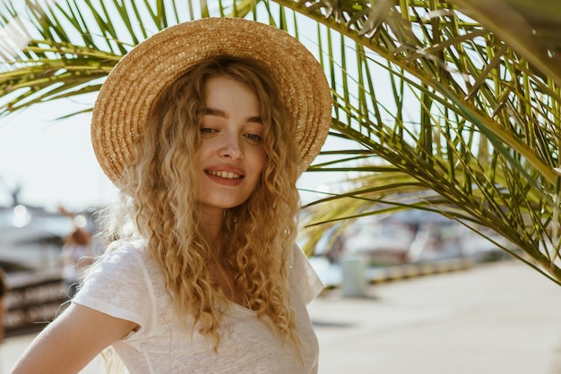 Curly blonde looking down gently smiles revealing beautiful teeth standing under a palm tree on the promenade