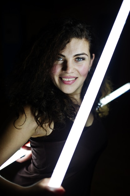 Curly brunette plus size model with two long led lamp at black room.