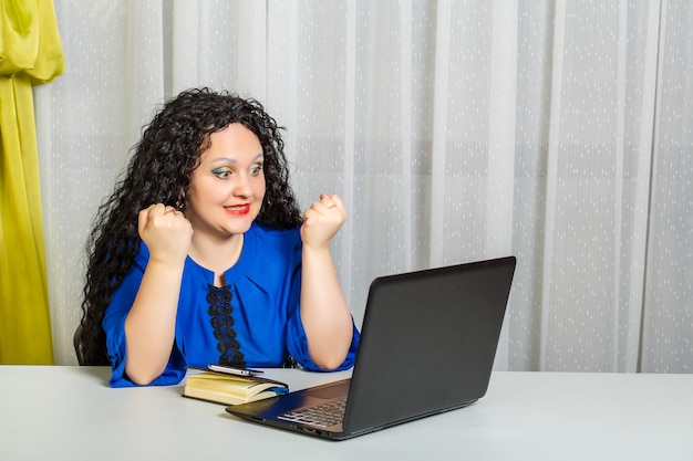 Curly brunette woman sits at a table in the office emotionally communicates via video communication. Horizontal photo