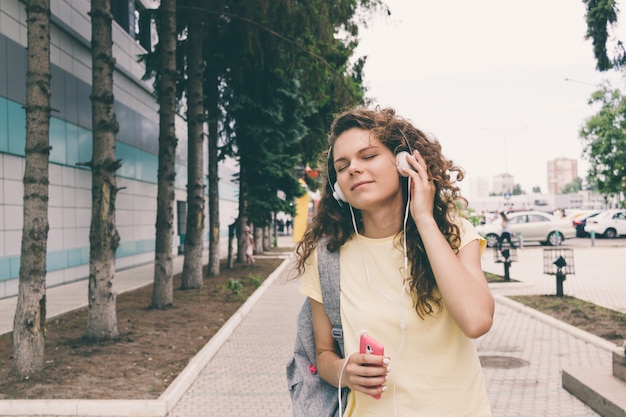 Curly brunette in a yellow T-shirt and headphones listening music walking around the city in summer