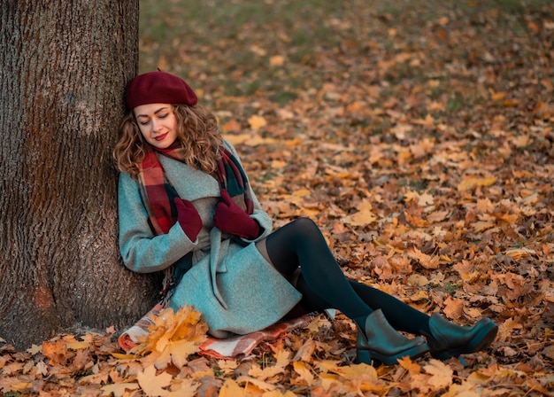 Curly cheerful girl with beautiful eyes wearing trendy gray coat and checkered scarf sitting near tree in the park.