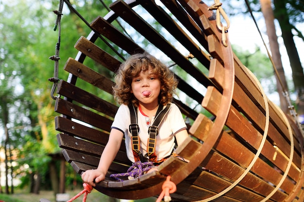 A curly child in climbing safety equipment in a tree house or in a rope park climbs the rope. 