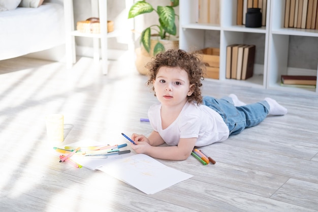 curly child girl drawing with colored markers lying on floor in living room at home