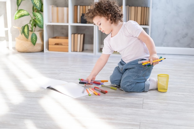 Curly child girl drawing with colored markers sitting on floor in living room at home