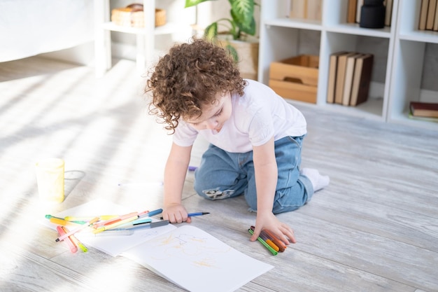 Curly child girl drawing with colored markers sitting on floor in living room at home