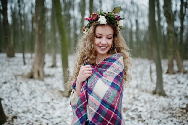 Curly cute blonde girl with wreath in checkered plaid at snowy forest in winter day.