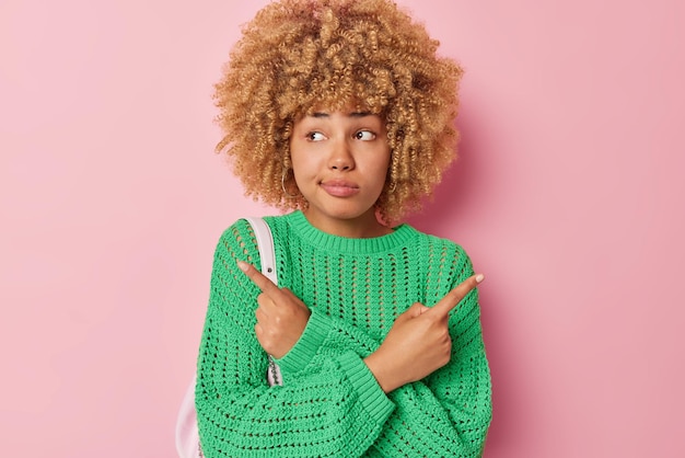 Curly doubtful curly woman points sideways shows left and right
product banner looks troubled by decision wears green knitted
sweater isolated over pink background needs help with right
choice