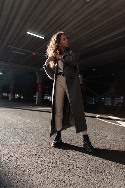 Curly fashion model girl in stylish vintage long coat, sweater, pants, boots and bag walks on the street. Urban female style and beauty. Sunlight and shadows