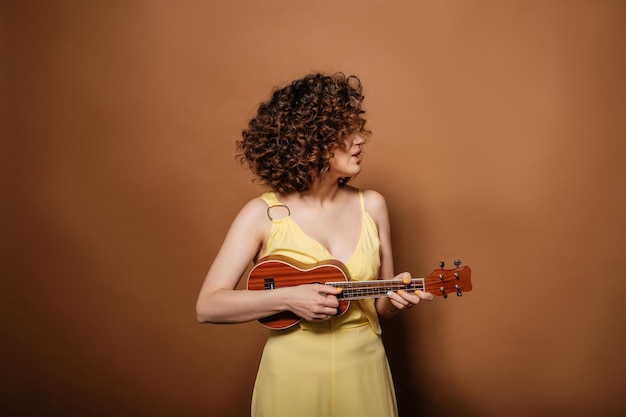 Photo curly girl in a yellow dress plays ukulele and sings a song