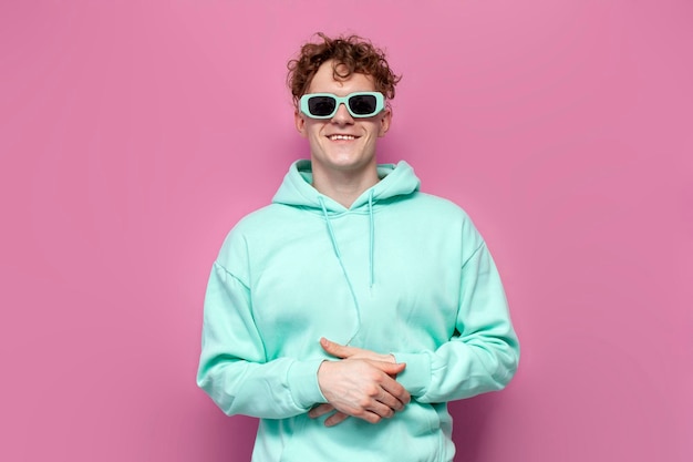 curly guy in mint hoodie and sunglasses smiles on pink isolated background man in blue sweatshirt