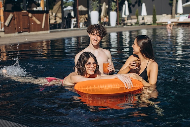 Curly guy and two beautiful girls drinking cocktails and having fun by the pool in summer