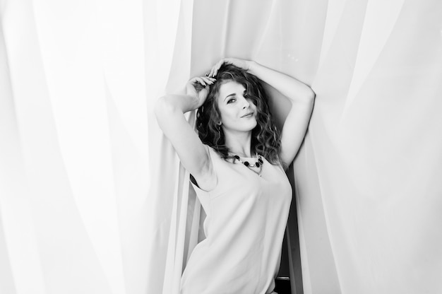 Curly hair girl on beige dress posed at white curtains.