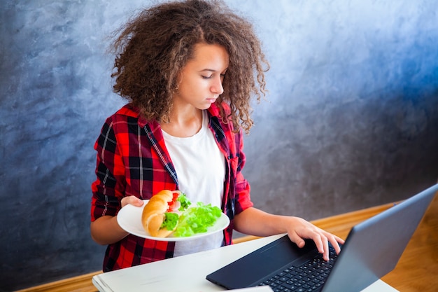 巻き毛の女の子のラップトップに取り組んでいるとサンドイッチを食べる