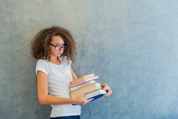 La ragazza teenager dei capelli ricci con i vetri sta accanto al muro e tiene parecchi libri