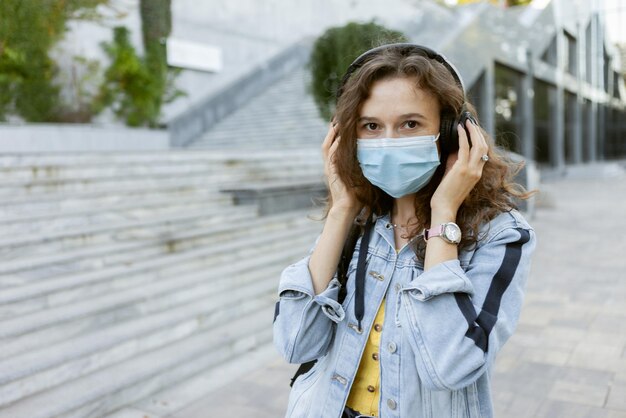 Curly hair woman in facial medical mask listens to music with headphones outdoors