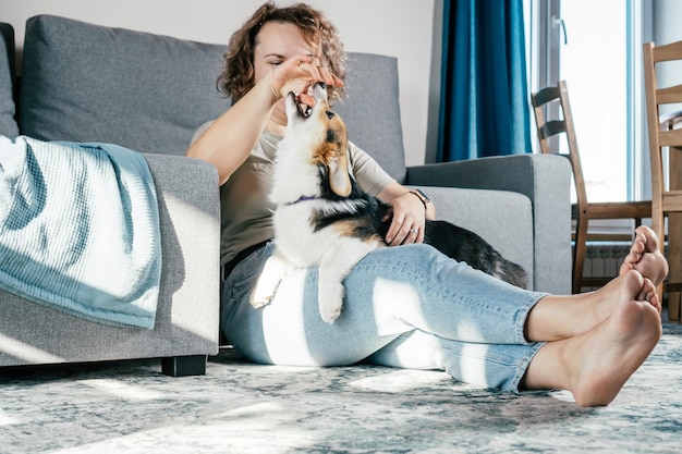 Curly haired blij vrolijke blote voeten vrouw met hond corgi gehoorzaamheid opleiding voeding van huisdier zittend ontspannen en spelen op de vloer in comfortabele woonkamer in de buurt van sofa Weekend vrijetijdsbesteding