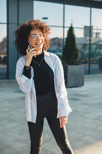 Curly haired business person talking on phone while smiling and wearing glasses outside