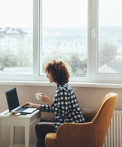 Curly haired caucasian woman dressed in casual clothes is drinking a coffee with sandwich while working at the laptop