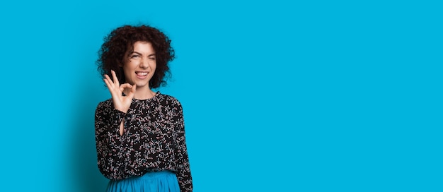 Curly haired  girl is gesturing the okay sign while posing near blue free space