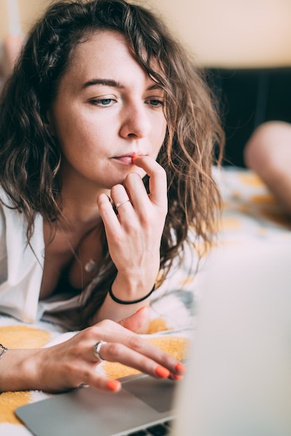 A curly-haired girl lies on the bed next to a laptop at home and with a serious face reads the news of the world.