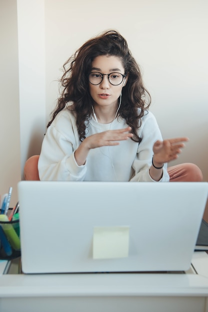 Curly haired jonge vrouw met bril is iets uit te leggen op de laptop tijdens een onlinevergadering vanuit huis