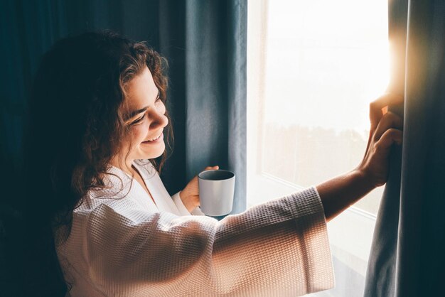 Photo curly haired lady in white bathrobe opens curtains to look outside large window drinking hot coffee