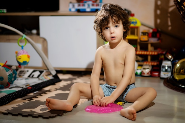 Curly-haired little boy in shorts plays with slime in his room, so as not to get his clothes dirty.