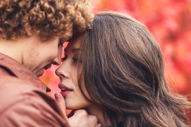 Curly-haired mustachioed man and brown-haired woman hugging in autumn against of red trees