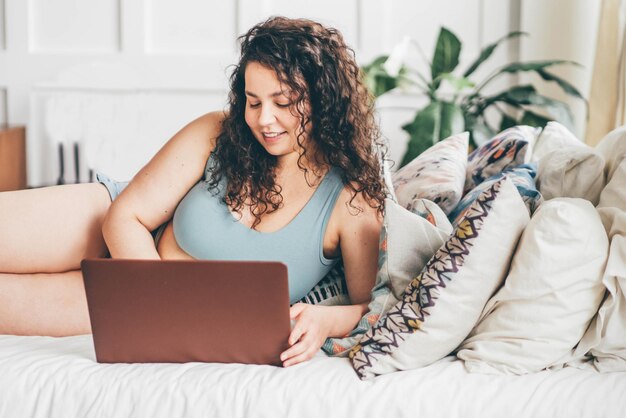 Curly haired overweight young woman in top and shorts types on\
laptop lying on queen size bed standing by window against wall in\
stylish light room