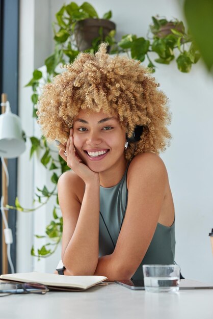 Curly haired schoolgirl listens audio course via stereo\
headphones writes down notes in notebook holds pen works on online\
project poses at desktop against cozy interior does remote distance\
job