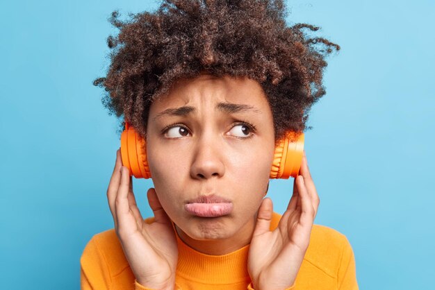 Curly haired upset woman listens audio song via wireless\
headphones frowns face purses lips wears orange jumper looks\
unhappily away isolated over blue background negative emotions\
concept