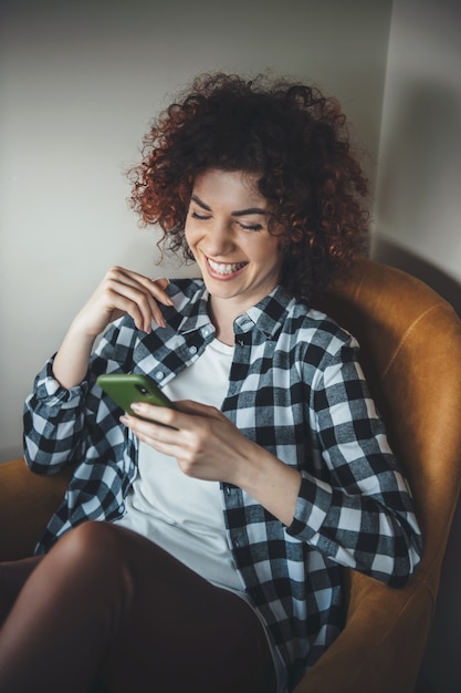 Curly haired vrouw die lacht in een fauteuil is sms'en op de telefoon thuis tijdens het op afstand onderwijs