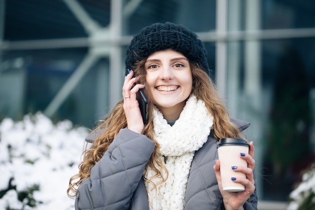 Curly haired vrouw met behulp van haar smartphone en papier beker met koffie te houden.
