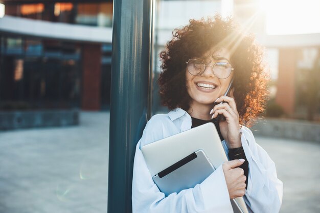 Curly haired vrouw praat over telefoon poseren buiten met een laptop bril