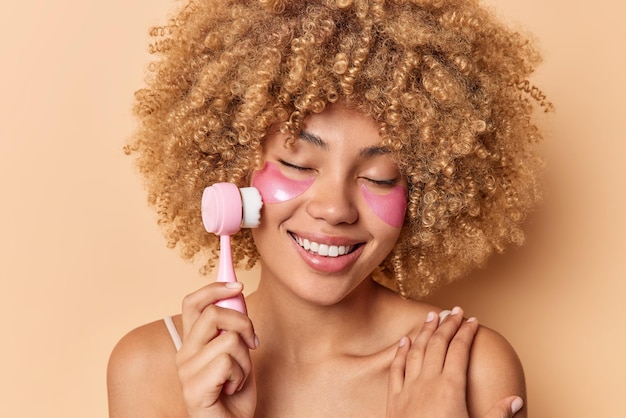 Curly haired woman uses face massager for skin treatment smiles happily applies pink hydrogel patches touches shoulders gently keeps eyes closed isolated over brown background. Beauty concept